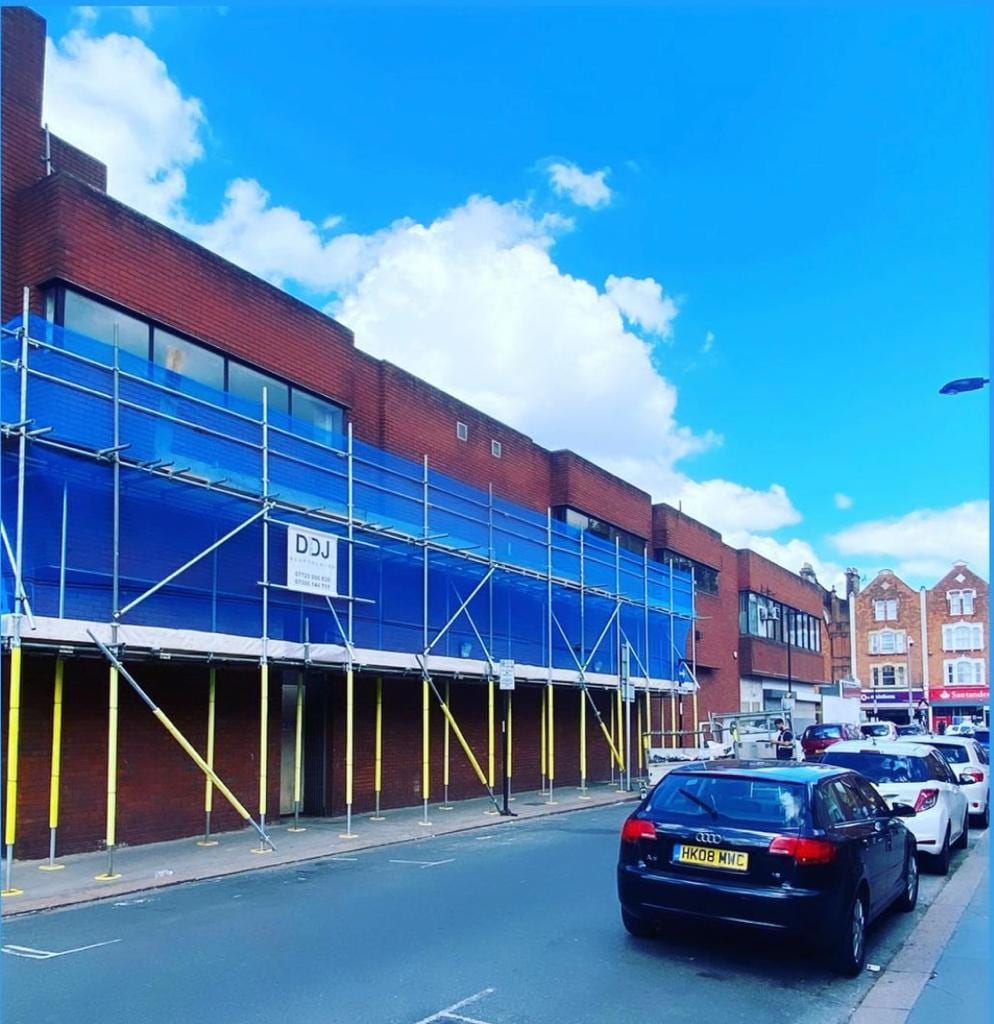 A long scaffold run along the glazed frontage of a retail parade beside a main road