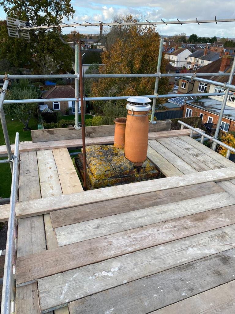 A boarded scaffold platform built around a chimney stack at roof level, guardrailed, with rooftops beyond
