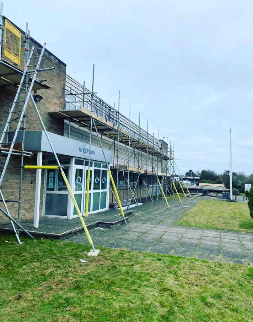 A long scaffold run along the frontage of a low commercial building beside its reception entrance, braced with raking props