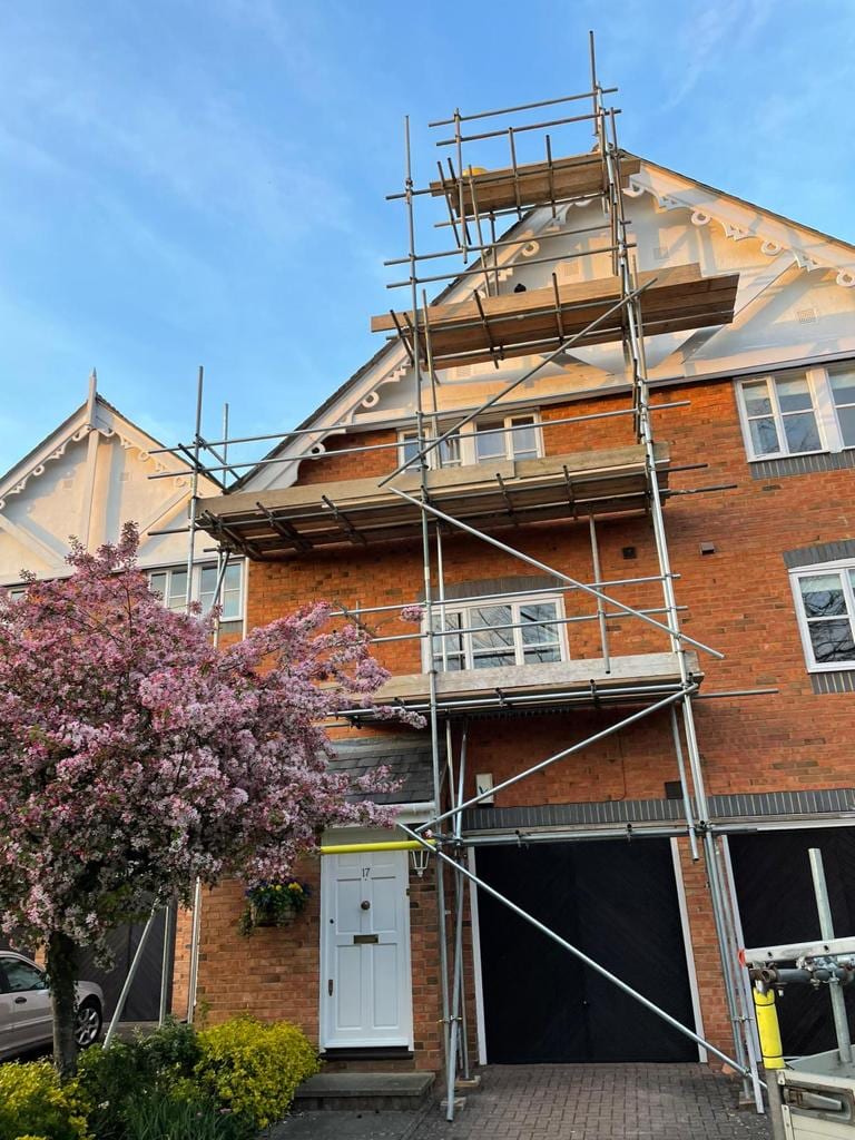 Scaffolding up the front of a tall brick townhouse beside a tree in blossom, boarded at two lifts