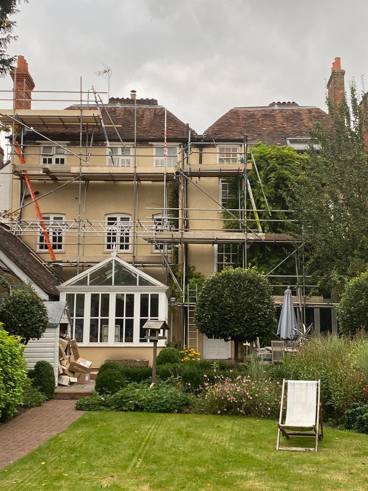 Three lifts of scaffolding across the rear of a large period house, above a conservatory and a planted garden