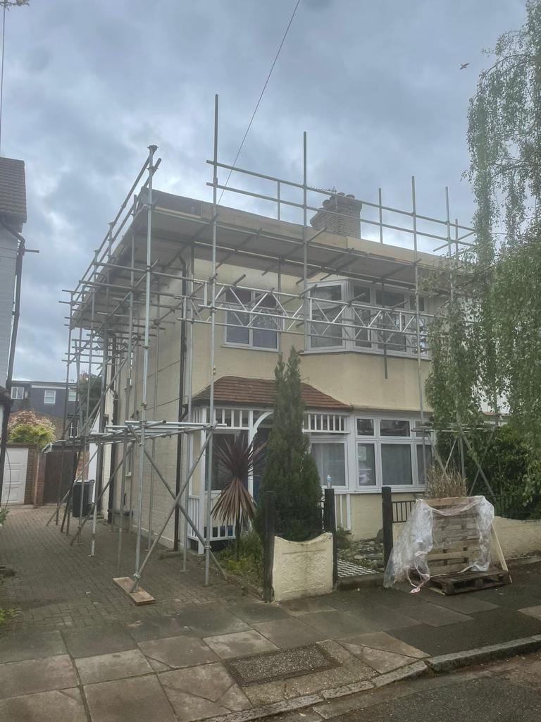 Scaffolding wrapped around the front and side of a rendered semi-detached house, seen from the pavement