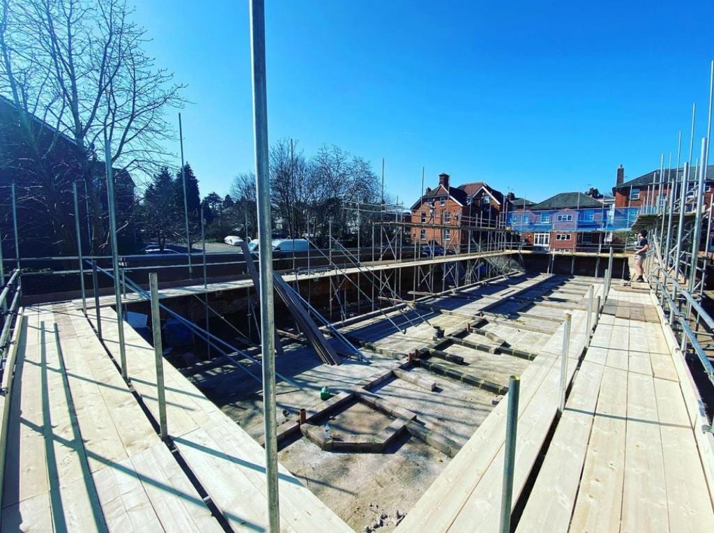 A fully boarded scaffold deck spanning an opened roof, with standards running up and rooftops beyond