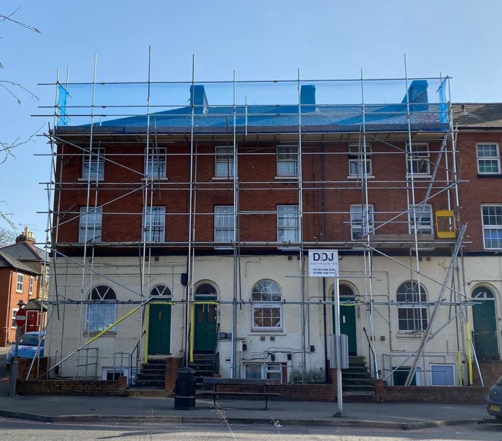 Full-height scaffolding across a three-storey brick terrace with arched doorways, netted at the top lift and carrying a DDJ Scaffolding board