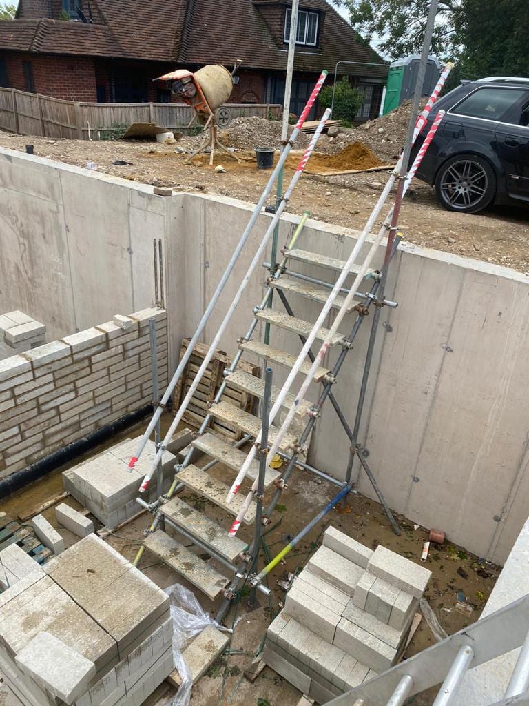 A scaffold stair tower running down into an excavated basement lined with concrete retaining walls, with stacked blocks alongside