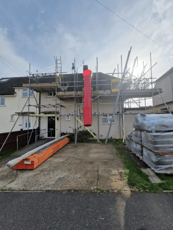 Scaffolding across the front of a rendered house with a debris chute hanging from the working lift, a timber ramp to the door and packs of boards stacked on the drive