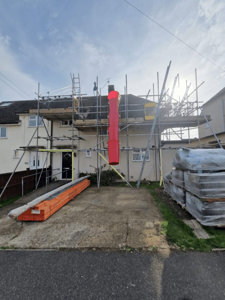 Scaffolding across the front of a rendered house with a debris chute hanging from the working lift, a timber ramp to the door and packs of boards stacked on the drive