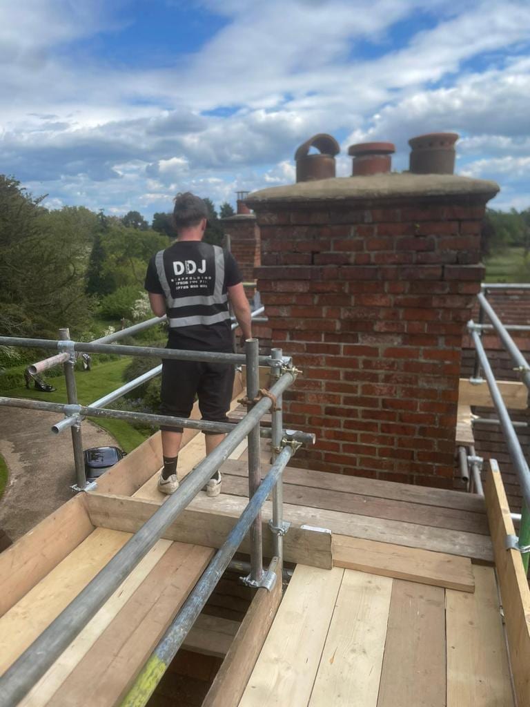 A scaffolder in DDJ branded workwear on a boarded platform beside a tall brick chimney stack with four pots, open country beyond