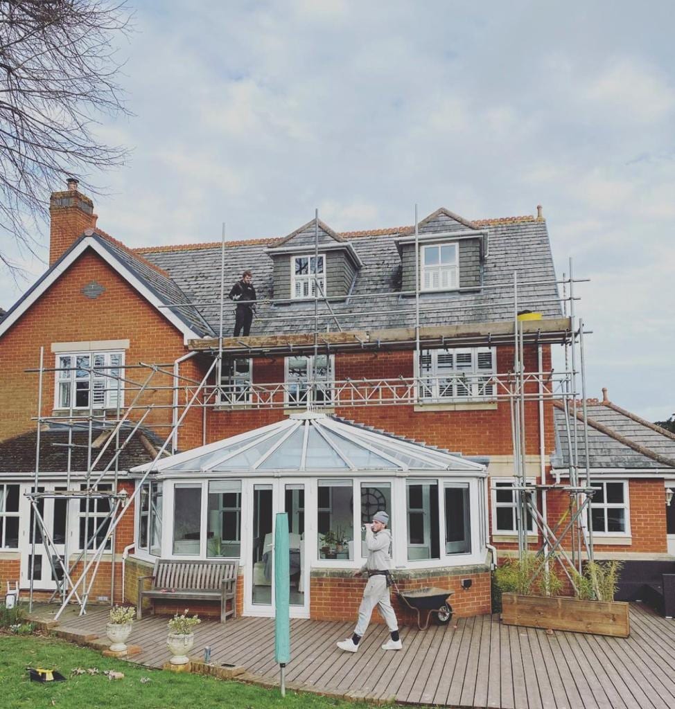 A scaffolder standing on a roof-level platform beside dormer windows, with the scaffold running above a conservatory and decking