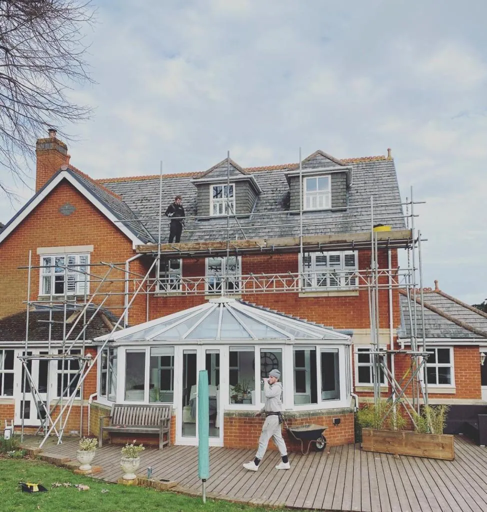 A scaffolder standing on a roof-level platform beside dormer windows, with the scaffold running above a conservatory and decking