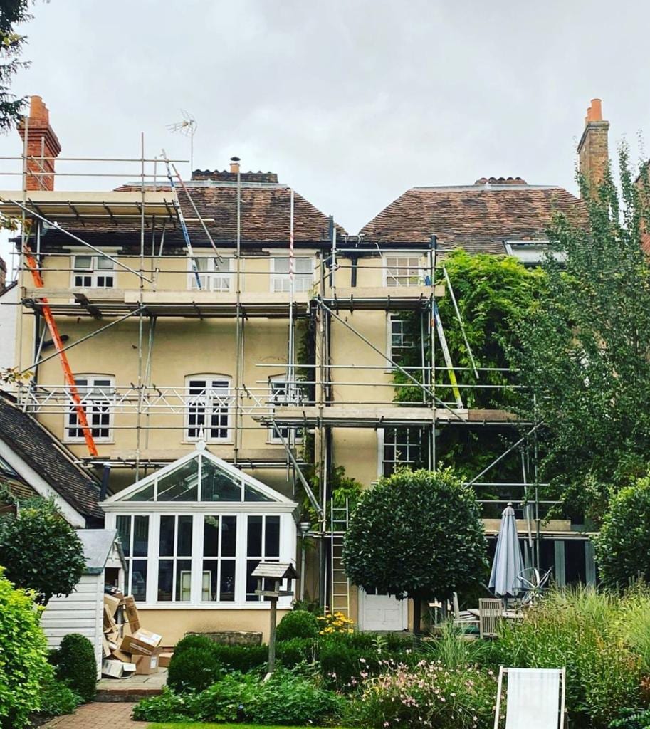 Two lifts of scaffolding across the rear of a large rendered period house, above a conservatory and a mature garden