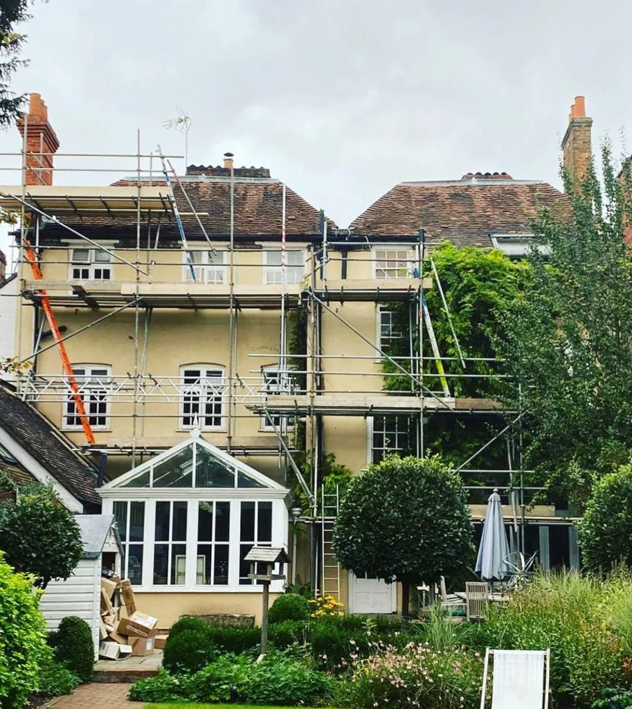 Two lifts of scaffolding across the rear of a large rendered period house, above a conservatory and a mature garden