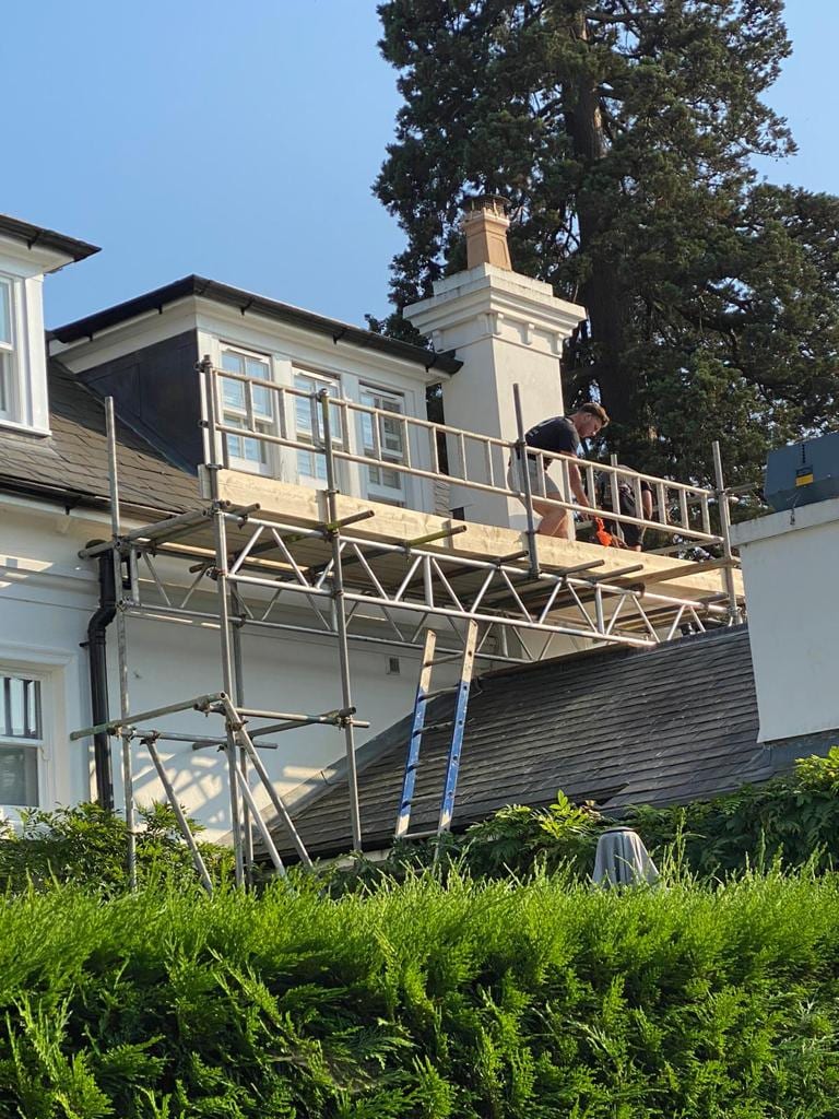 A scaffolder working from a guarded platform beside a tall rendered chimney stack, level with the roof of a white house