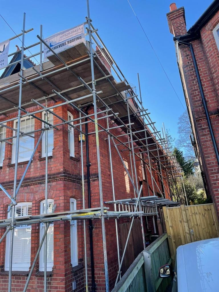 Scaffolding rising around the corner of a Victorian brick house, boarded and guarded at eaves level