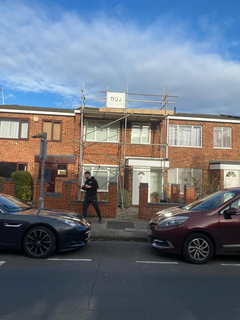 A single-bay scaffold up the front of a brick mid-terrace house, with a scaffolder walking past on the pavement