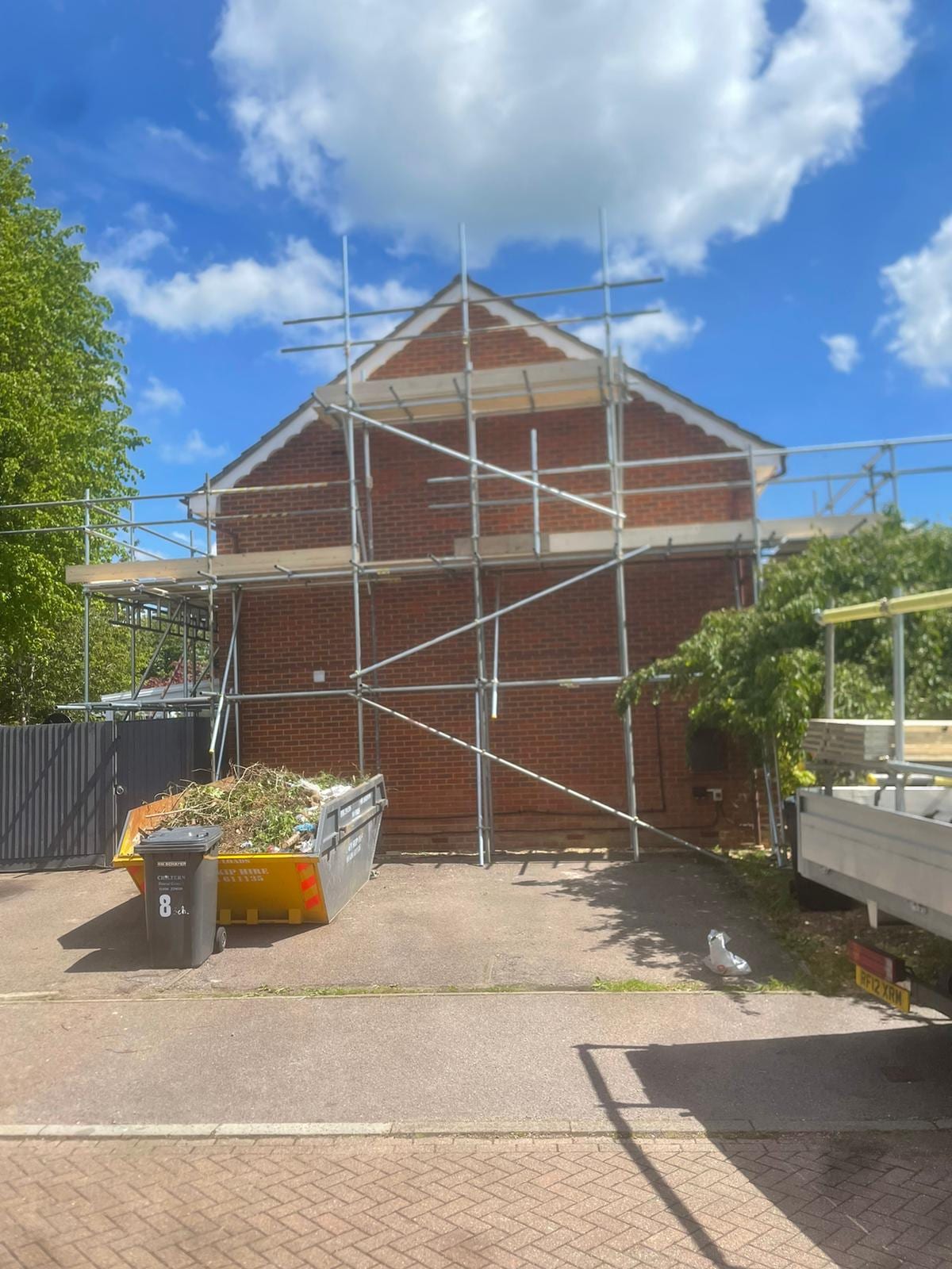 Scaffolding wrapped around the gable end of a brick house beside a driveway and a loaded skip