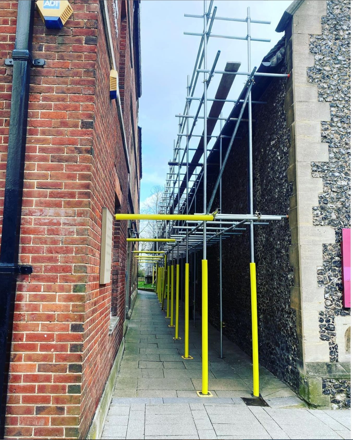 A scaffold run threaded down a narrow alleyway between a brick building and a flint wall, braced to the paving