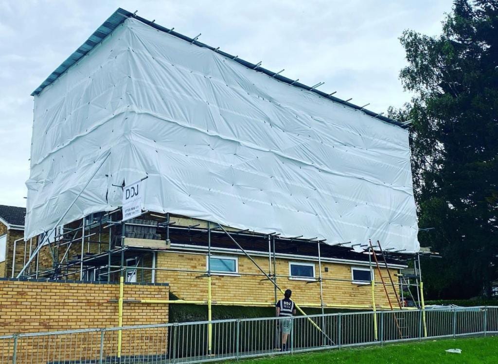 A low building entirely encapsulated in shrink-wrap sheeting on scaffold, with a DDJ scaffolder standing at the boundary rail
