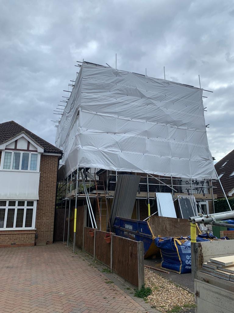 The upper storey of a house wrapped in sheeting on a scaffold, with a skip and stacked sheets on the drive below