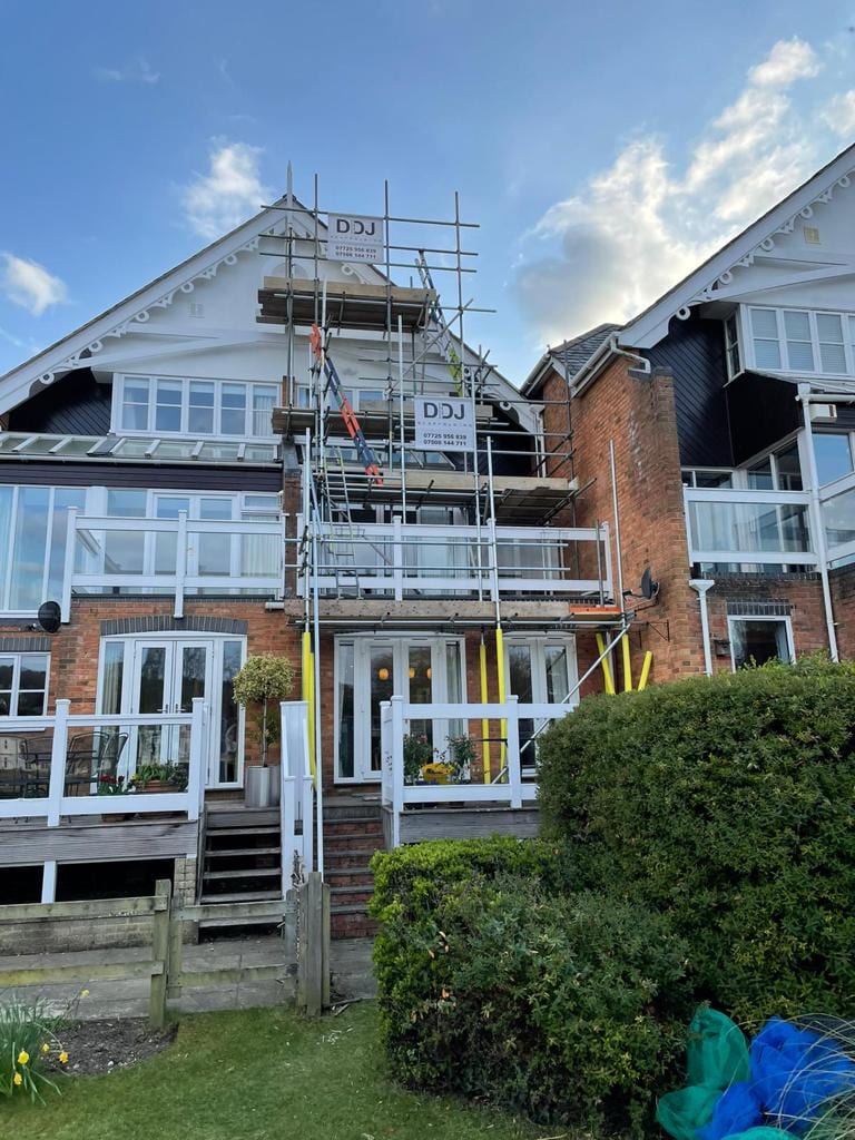 Scaffolding up the front of a large gabled house with balconies, carrying a DDJ Scaffolding banner